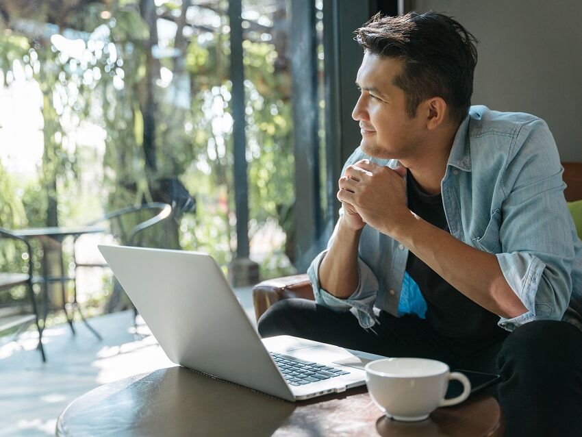 Man looking out window with laptop and coffee cup on table in front of him