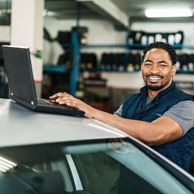 Male mechanic typing on computer inside of garage with car in foreground
