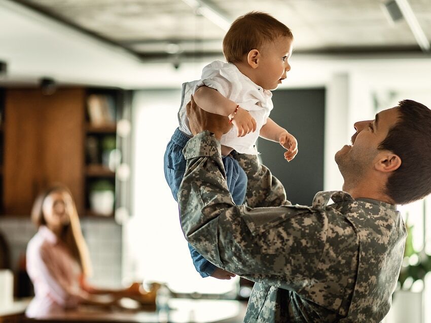 Man in military uniform holding baby boy in kitchen with woman in the background.