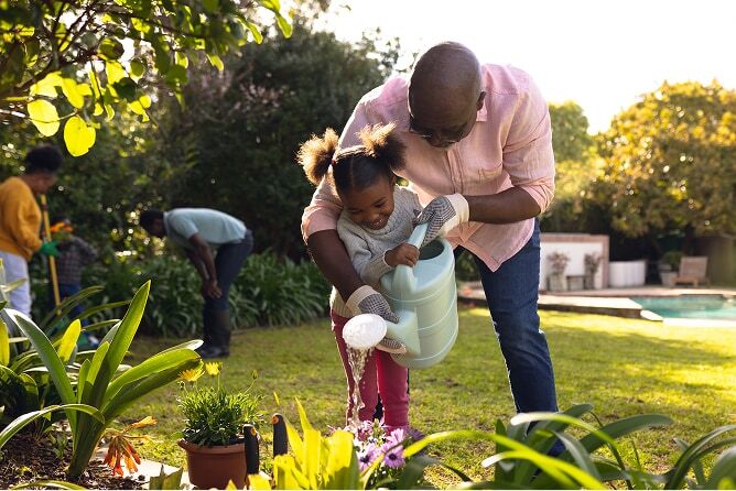Father helping daughter water flower garden with other family members in the background