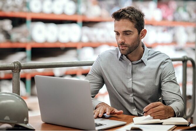 Man working on laptop with papers on table and warehouse shelves in background