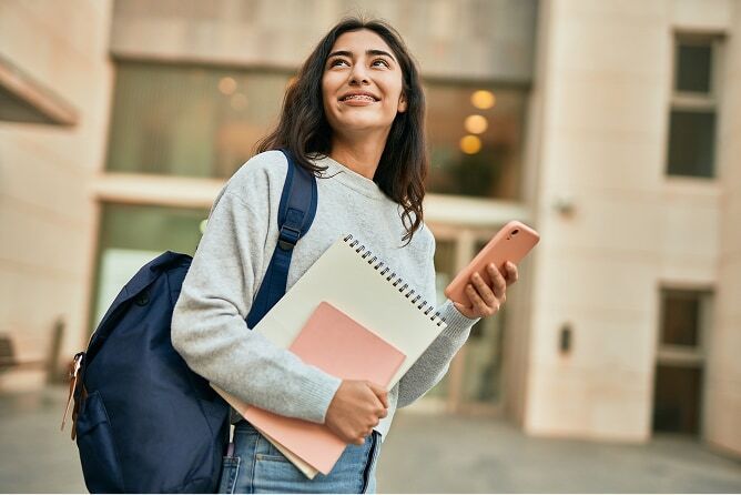Young college student holding books, phone, and backpack in front of campus building