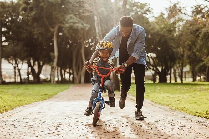 Father teaching son to ride a bike on brick sidewalk with trees in background