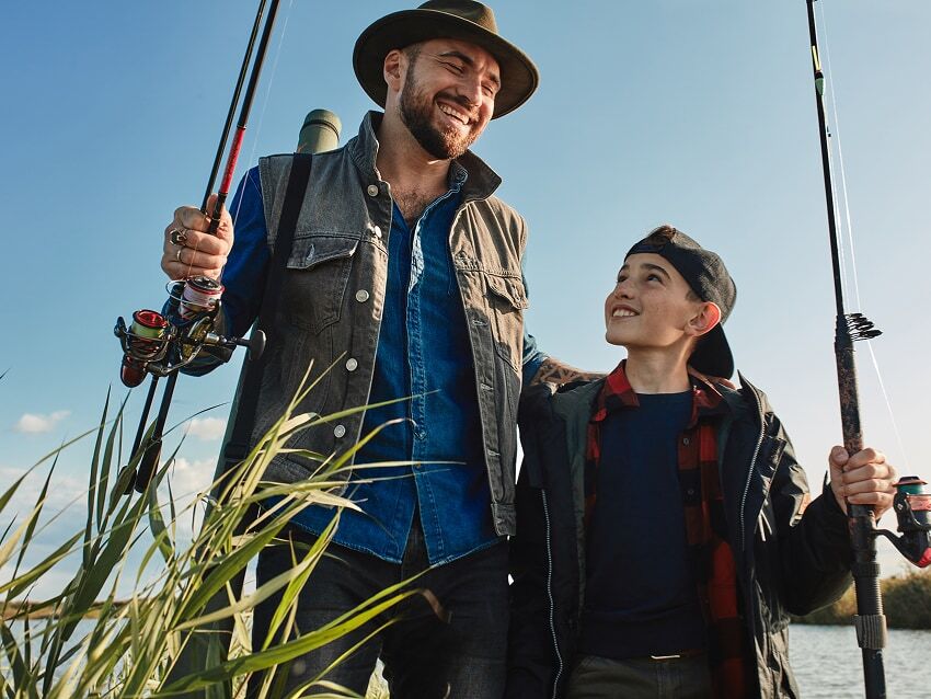 smiling father and son walking through tall grass carrying fishing poles