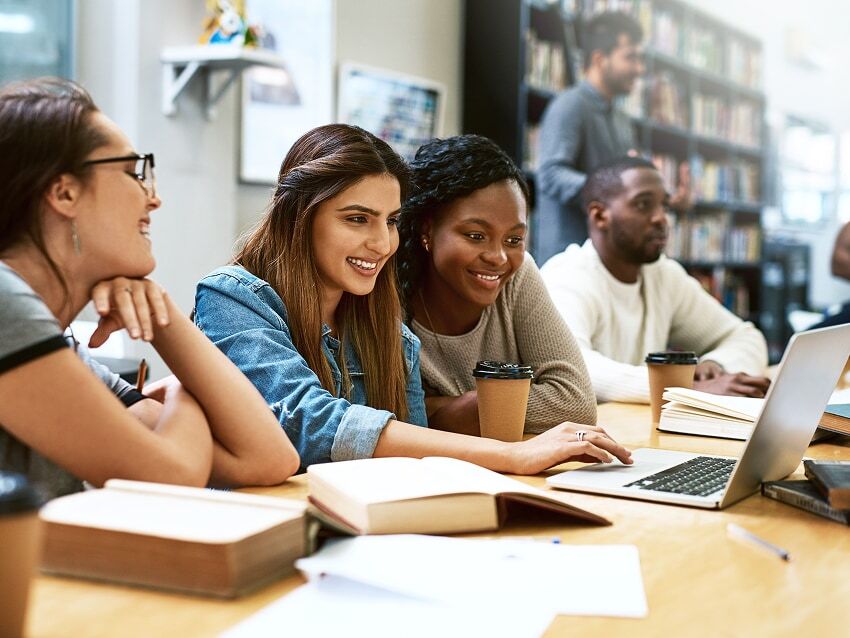 Group of students at a library table with books and laptop