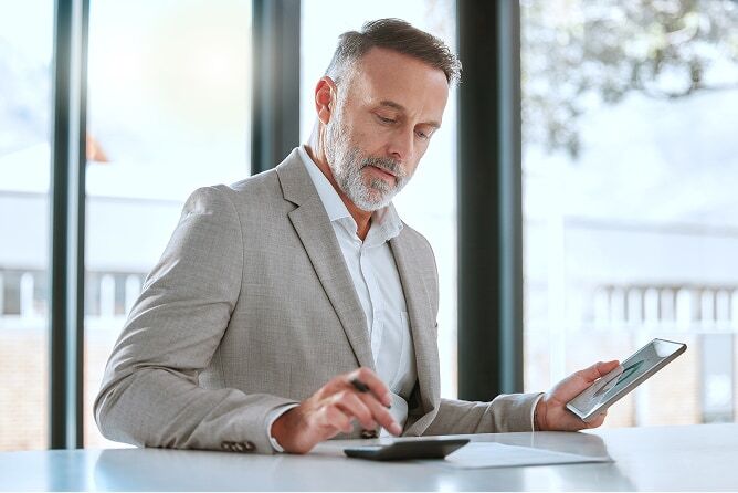 Businessman in suit reviewing digital financial report and writing on piece of paper