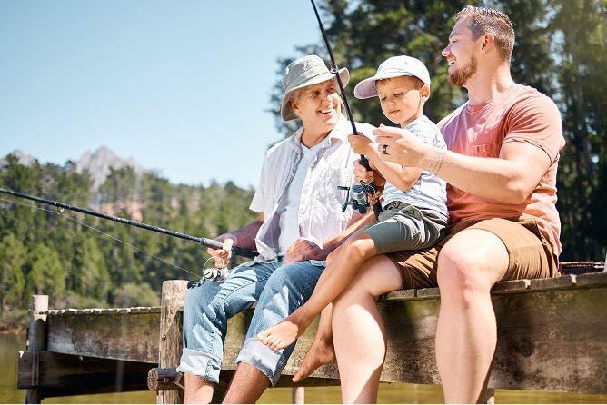Grandfather, father, and young child sitting on dock fishing with mountains in background