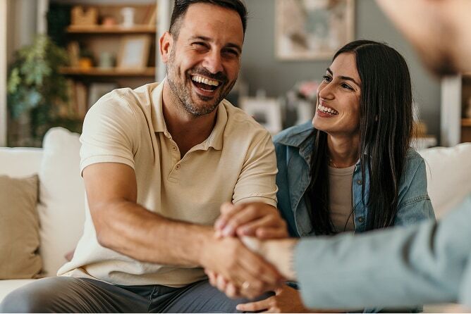 Husband shaking hands with advisor with smiling wife beside him on couch